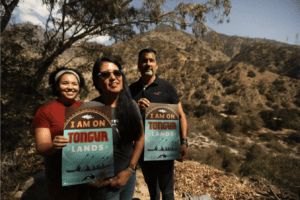A photo of three people on a chaparral-covered hillseide holding "you are on tongva land" signs