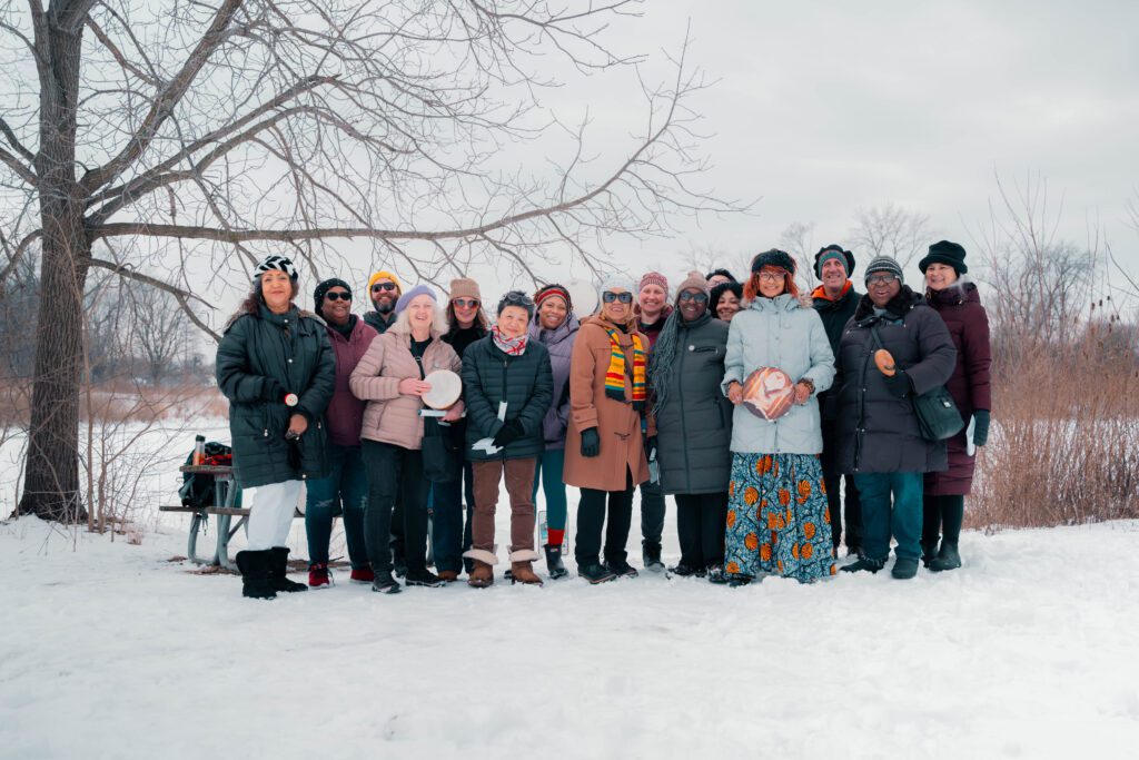 A group picture of 16 people at "Down by the River" with the snowy Milwaukee River behind them